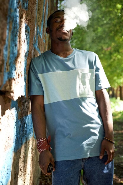 Portrait of African American guy in casualwear smoking by wall of building