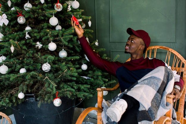 Portrait of an African-American hipster guy dressed in a red fleece sweater and red cap sitting in the a comfortable chair near the Christmas tree