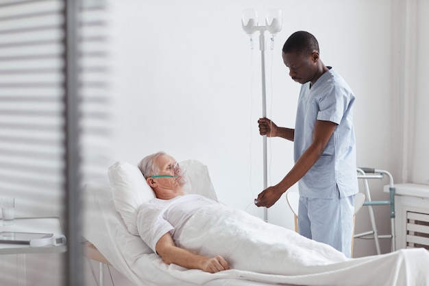 Ritratto di un infermiere afroamericano che prepara flebo iv mentre tratta un paziente anziano nel letto d'ospedale, copia spazio