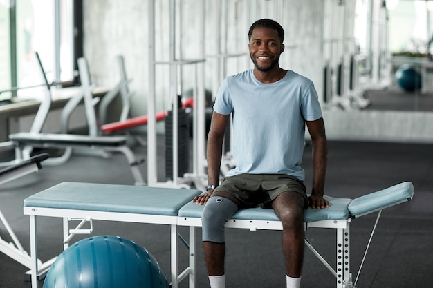 Portrait of african american man sitting on massage bed in\
rehabilitation clinic and smiling at camera