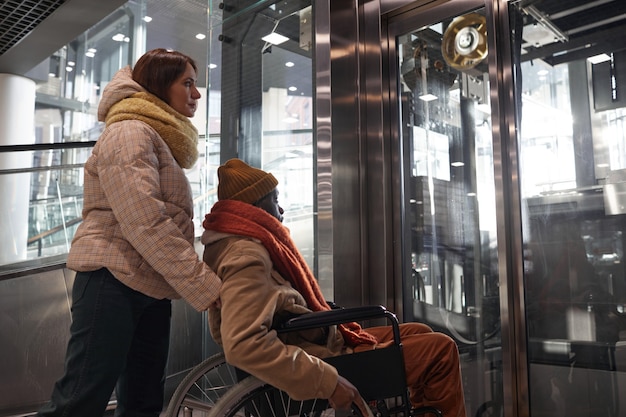 Portrait of African American man in wheelchair using accessible elevator with young woman assisting in urban city setting, copy space