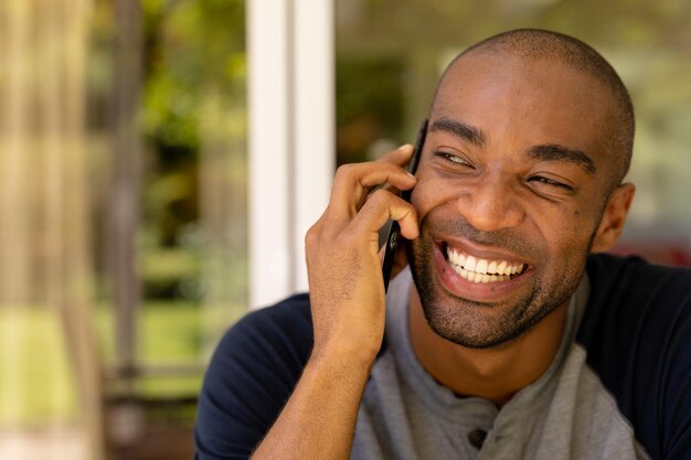 Portrait of an African American man with gray t shirt sitting in the patio, talking on his mobile phone and laughing during a sunny day