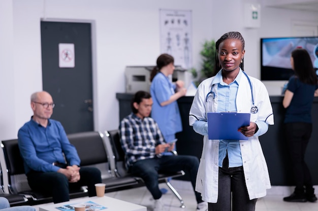 Ritratto di medico afroamericano in posa fiducioso nel camice da laboratorio e in possesso di appunti in attesa del prossimo appuntamento del paziente. medico sorridente con stetoscopio in un'affollata reception dell'ospedale.