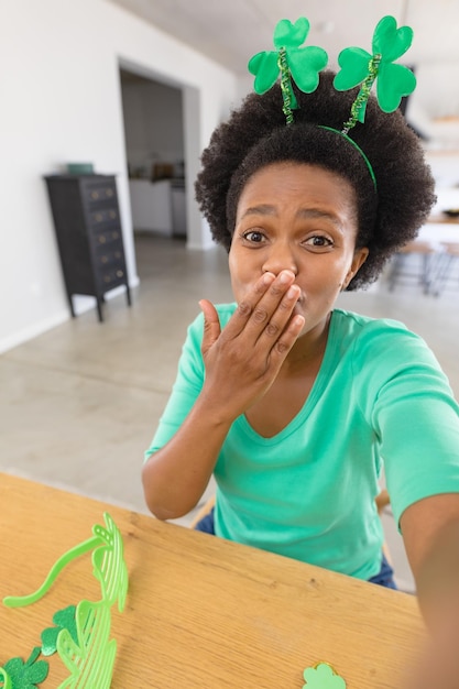 Portrait of african american mid adult woman in shamrock headband blowing kiss on video call. unaltered, lifestyle, st patrick's day, holiday and celebration concept.