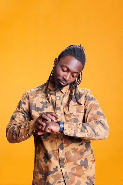 Portrait of african american person checking time on wristwatch, being late at work. Young adult worrying about punctuality, looking at smartwatch in studio over yellow background.