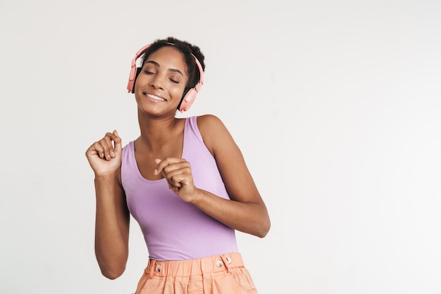 Portrait of african american positive woman using headphones and dancing isolated on white