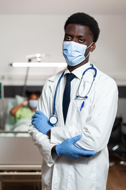 Portrait of african american practitioner doctor with protective face mask to prevent infection with covid19 looking into camera while standing in hospital ward. In background patient recovering