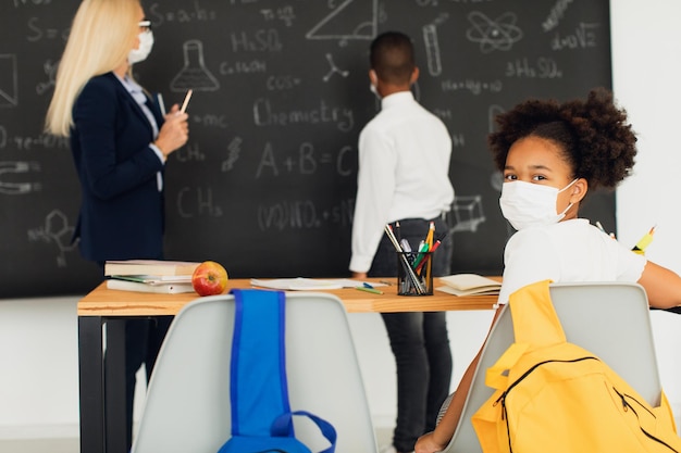 Portrait of african american school girl in mask sitting at\
desk on a blackboard background in a classroom during a lesson mask\
mode quarantine covid19