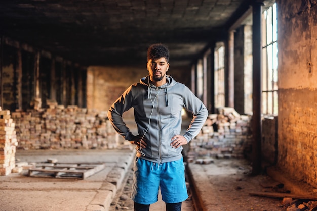 Portrait of african american sporty man with serious face holding hands on hips and standing in old brick factory.