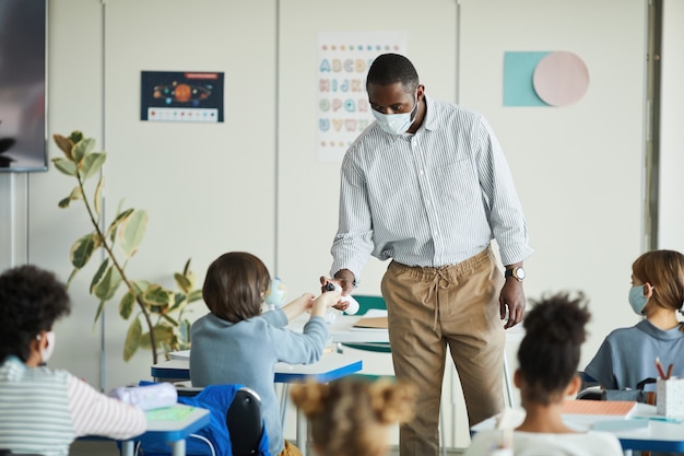 Photo portrait of african-american teacher sanitizing hands of children in school classroom, covid safety measures, copy space
