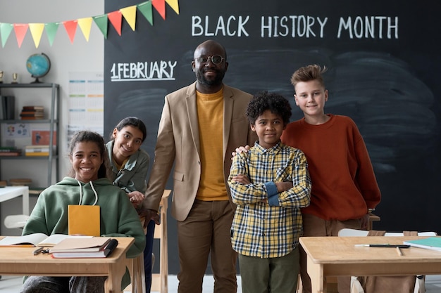 Photo portrait of african american teacher smiling at camera together with his pupils while they standing in classroom against blackboard