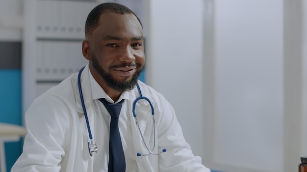 Portrait of african american therapist doctor sitting at desk in hospital office analyzing sickness document on computer typing medical expertise. Physician man working at healthcare treatment