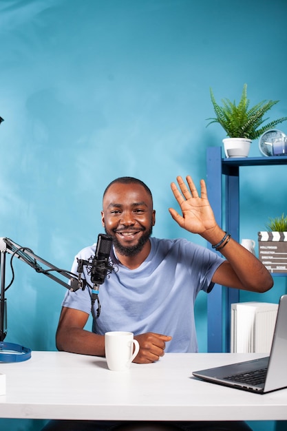 Portrait of african american vlogger waving hand at camera in\
recording studio with professional microphone and broadcasting\
setup. content creator saying hello at audience during online live\
show.