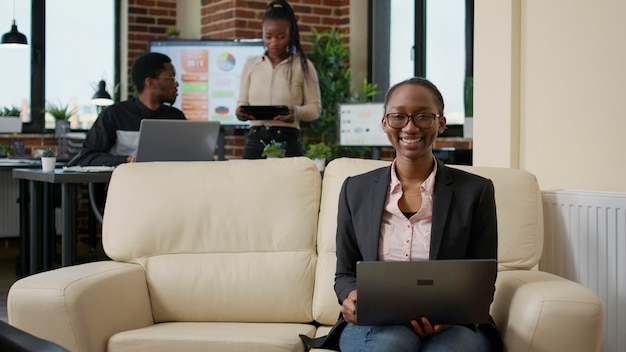 Ritratto di donna afroamericana che tiene laptop sul divano nell'ufficio aziendale, utilizzando la rete internet per analizzare le informazioni finanziarie per la crescita e lo sviluppo del business. carriera esecutiva.