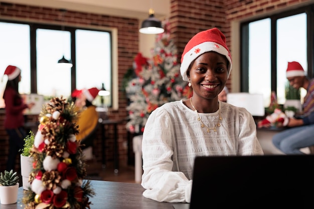 Portrait of african american woman with santa hat working on business at company office filled with christmas decorations and tree lights. Using laptop in startup workplace with xmas decor.