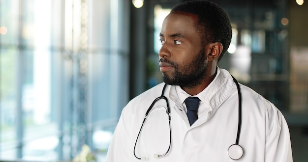 Portrait of African American young man doctor with stethoscope looking at camera and smiling cheerfully. Handsome male physician smile. Medic in white gown. Indoors.