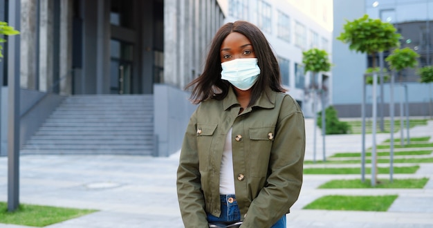 Ritratto di giovane donna graziosa alla moda afroamericana in mascherina medica che guarda l'obbiettivo e in piedi in strada urbana. bella femmina all'aperto in città durante la pandemia. confinamento per il coronavirus.