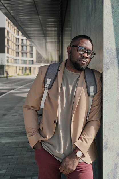 Portrait of African businessman in eyeglasses with backpack behind his back looking at camera while standing in the city