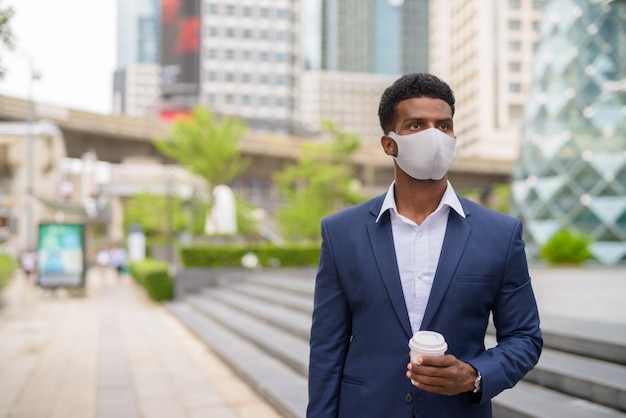 Portrait of African businessman wearing face mask outdoors in city and holding take away coffee cup, horizontal shot