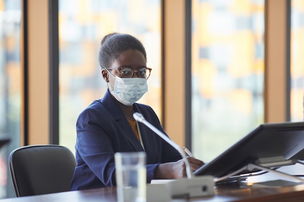 Portrait of African businesswoman in eyeglasses and in protective mask looking  while sitting at the table in board room