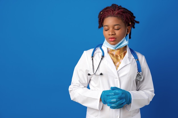 Portrait of african female doctor in lab coat with face mask and stethoscope against blue background