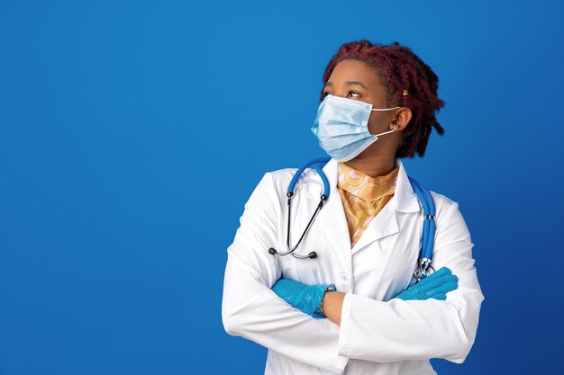 Portrait of african female doctor in lab coat with face mask and stethoscope against blue background