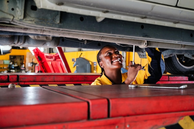 Foto ritratto di una meccanica africana in uniforme giallo e blu in piedi sotto il fondo dell'auto