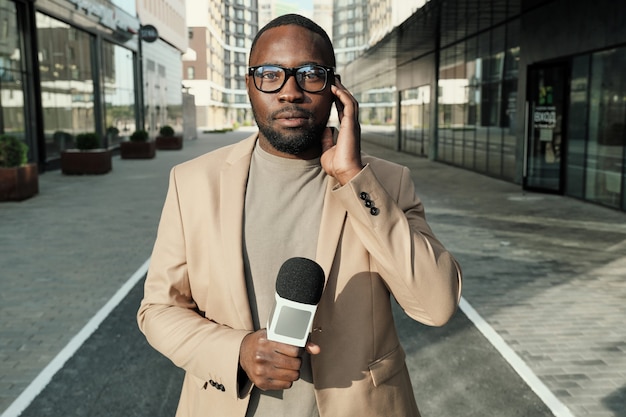 Portrait of African journalist standing with microphone and looking at camera during his work in the city