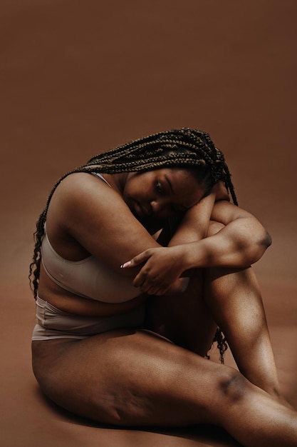 Portrait of african plump woman in underwear sitting against the brown background and looking very sad