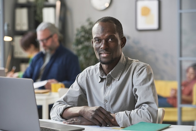 Portrait of African young businessman smiling at camera while sitting at the table in front of the laptop at office