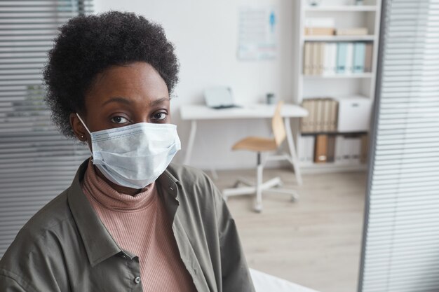 Portrait of African young woman in protective mask looking at camera while sitting at doctor's office