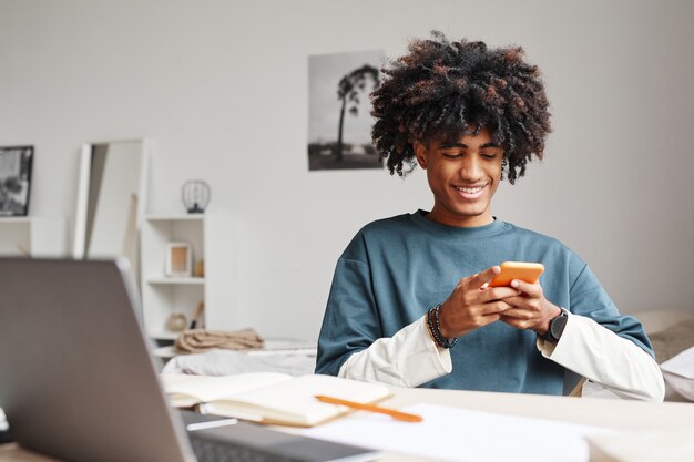 Portrait of africanamerican teenage boy using smartphone and smiling while studying at home or in co...