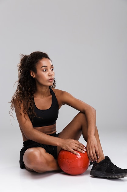 灰色の背景の上に分離された重いボール、座って、目をそらしてエクササイズをしているアフリカ系アメリカ人の自信を持って若いフィットスポーツウーマンの肖像画