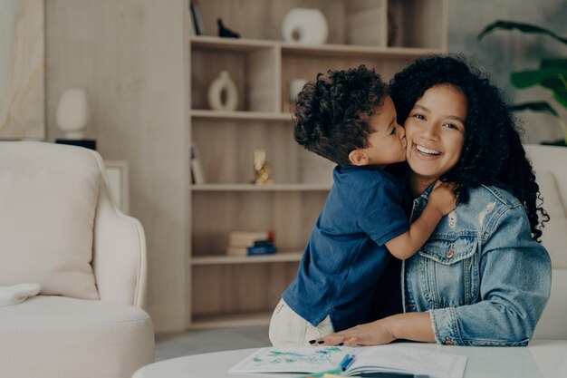 Photo portrait of afro american cute boy in blue polo tshirt hugging mother and kissing her in cheek