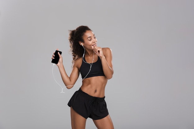 Portrait of an afro american happy sportswoman isolated over gray background, listening to music with earphones, holding mobile phone, dancing