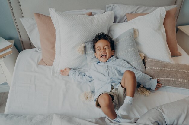 Portrait of afro american joyful little boy on pillows in bed laughing and smiling