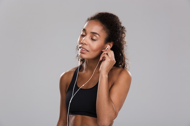 Портрет молодой афро-американской спортсменки, изолированной на сером фоне, слушающей музыку в наушниках с закрытыми глазами