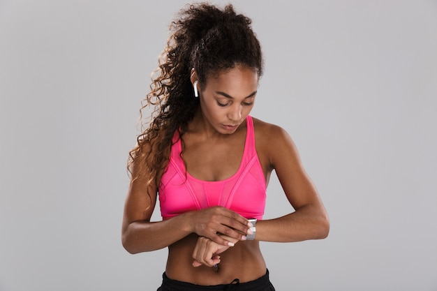 Portrait of an afro american young sportswoman isolated over gray background, listening to music with earphones, looking at her smart watch