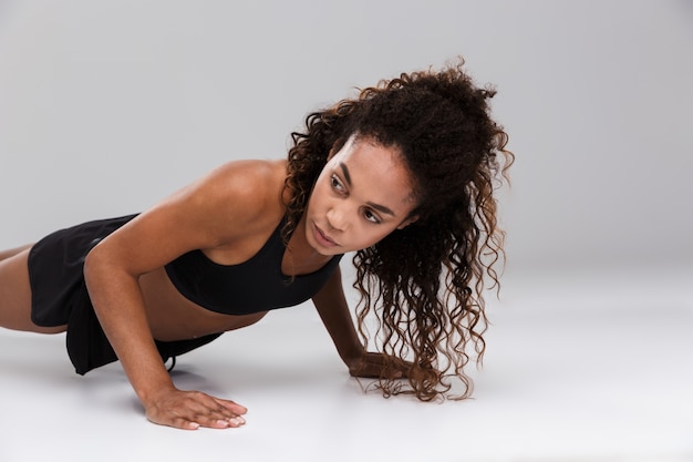 Portrait of an afro american young strong sportswoman doing plank exercise isolated over gray background
