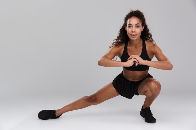 Portrait of an afro american young strong sportswoman isolated over gray background, doing side lunges