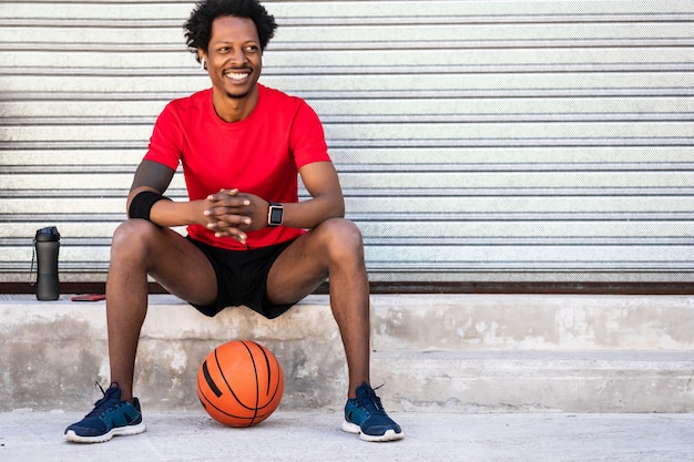 Portrait of afro athlete man holding a basketball ball and relaxing after training while sitting outdoors. Sport and healthy lifestyle.