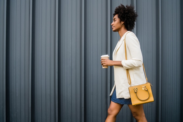 Portrait of afro business woman holding a cup of coffee while walking outdoors on the street. Business and urban concept.