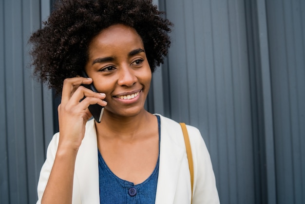 Portrait of afro business woman talking on the phone and holding a cup of coffee while standing outdoors at the street