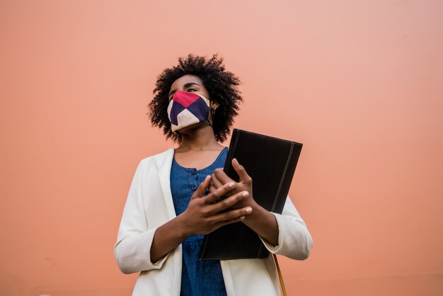 Ritratto di afro imprenditrice indossando maschera protettiva e tenendo un appunti mentre si trova all'aperto in strada