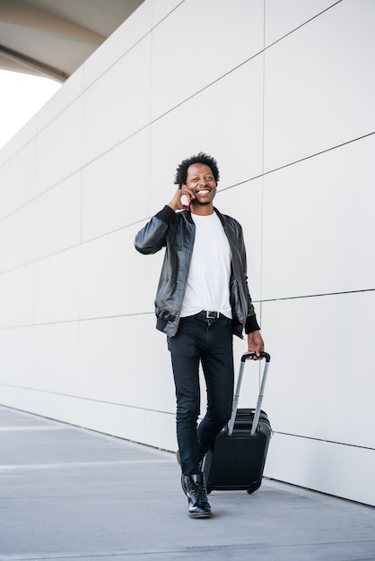 Portrait of afro tourist man talking on the phone and carrying suitcase while walking outdoors on the street. Tourism concept.