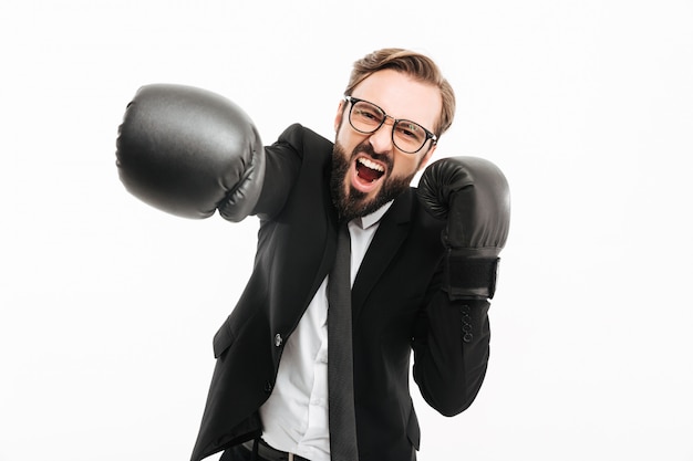 Portrait of aggressive businessman in black suit and eyeglasses screaming while punching in boxing gloves, isolated over white wall