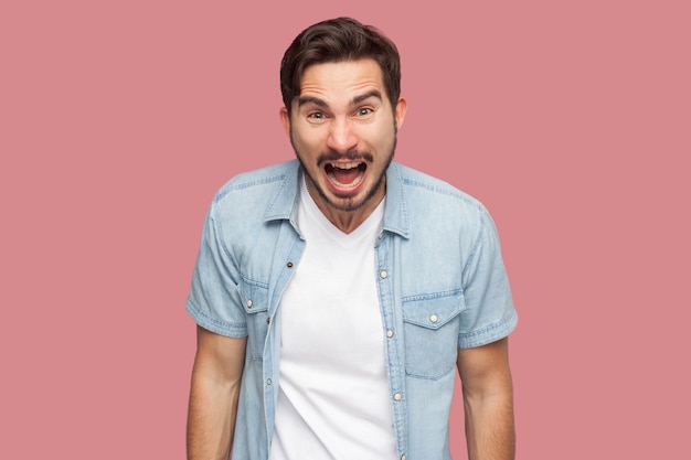 Portrait of aggressive shocked handsome bearded young man in blue casual style shirt standing looking and screaming at camera with angry face. indoor studio shot, isolated on pink background.