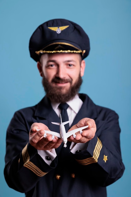 手のひらの正面図で飛行機のおもちゃを保持している制服を着た飛行機飛行士のポートレート、商用旅客機モデルで遊ぶパイロット。青の背景にプロの航空アカデミー飛行士