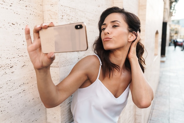 Portrait of alluring pleased woman making kiss face and taking selfie photo on smartphone while standing over wall on city street