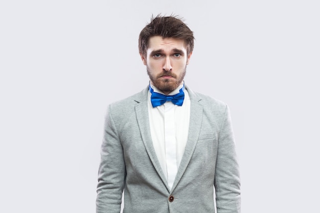 Portrait of alone handsome bearded man in casual grey suit and blue bow tie standing and looking at camera with sad eyes. indoor studio shot, isolated on light grey background.